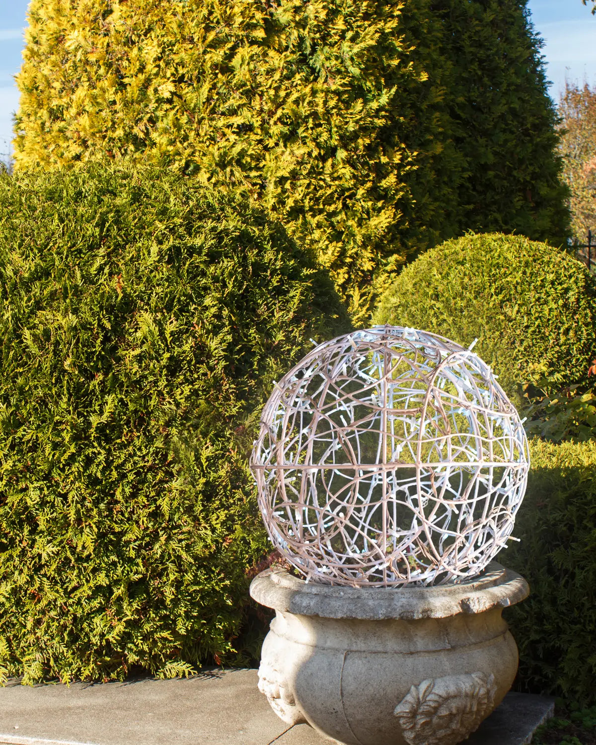 Weiße Lichtkugel aus Aluminiumgeflecht auf Steinsockel – dezente Tagesinstallation im Gartenbereich.