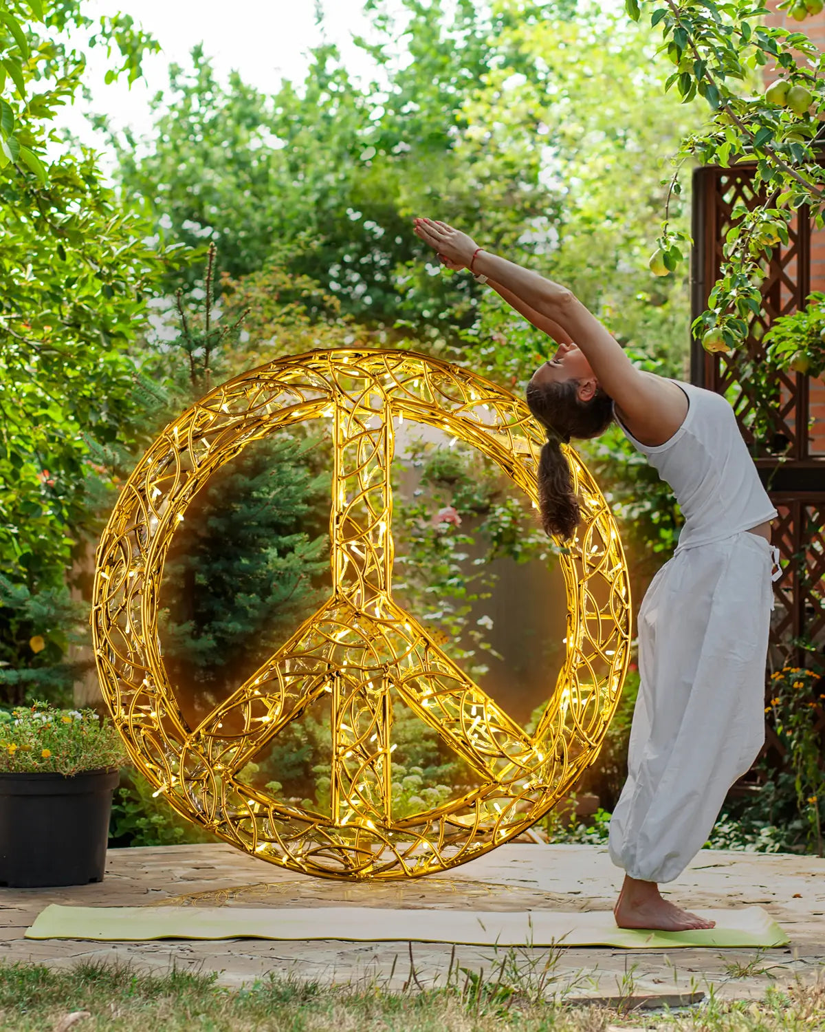 Frau macht Yoga-Übung im Garten vor einer leuchtenden Friedenssymbol-Skulptur aus goldenen Lichtdrähten, umgeben von grüner Natur.