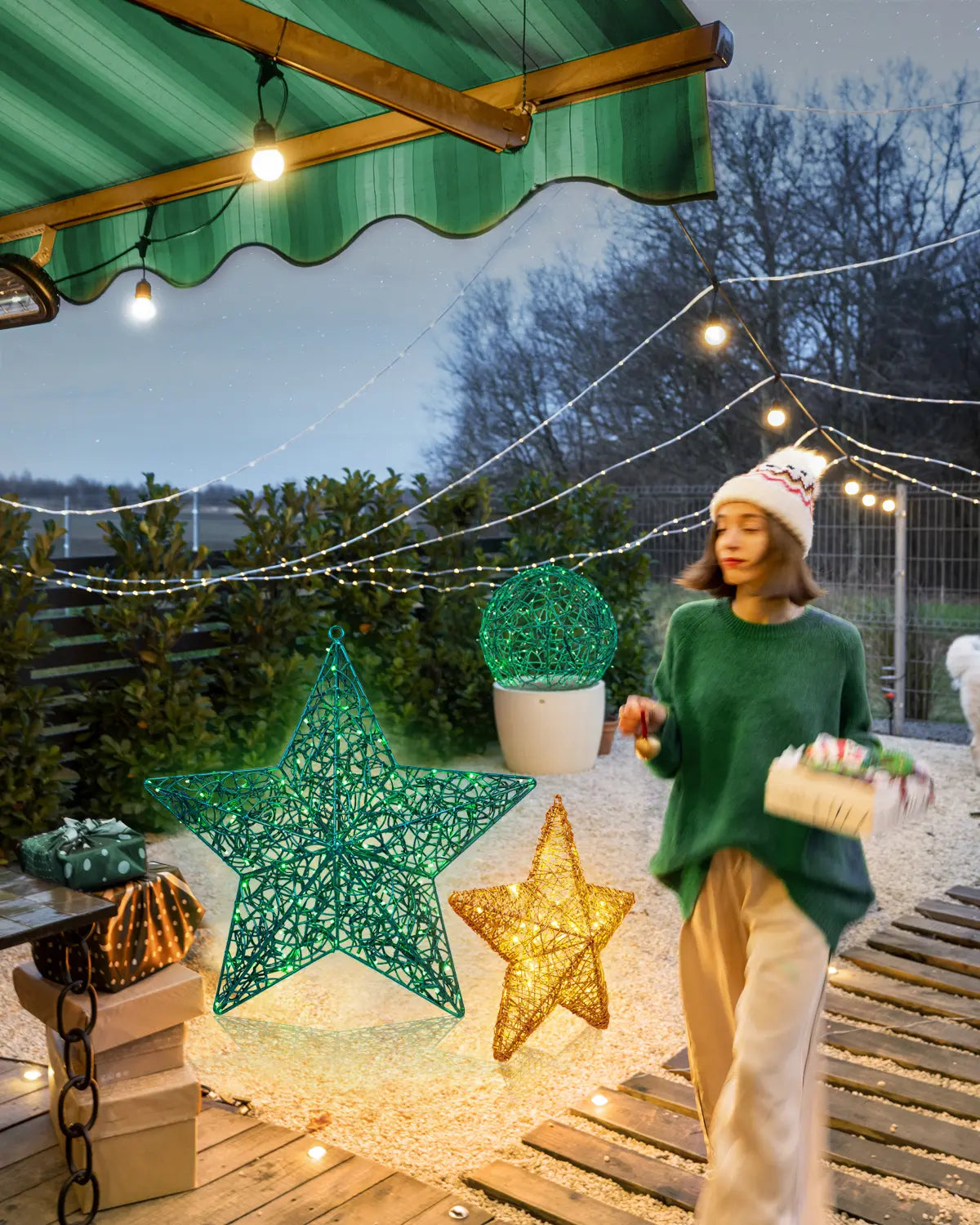 Außenszene mit grünen und goldenen Lichtobjekten in Stern- und Kugelform – winterlich dekorierter Gartenbereich mit Lichterketten und Geschenkübergabe.