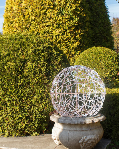 Weiße Lichtkugel aus Aluminiumgeflecht auf Steinsockel – dezente Tagesinstallation im Gartenbereich.