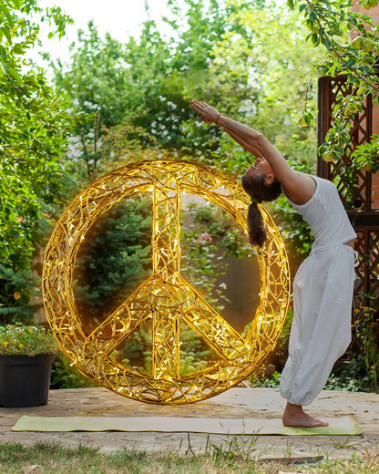 Frau macht Yoga-Übung im Garten vor einer leuchtenden Friedenssymbol-Skulptur aus goldenen Lichtdrähten, umgeben von grüner Natur.